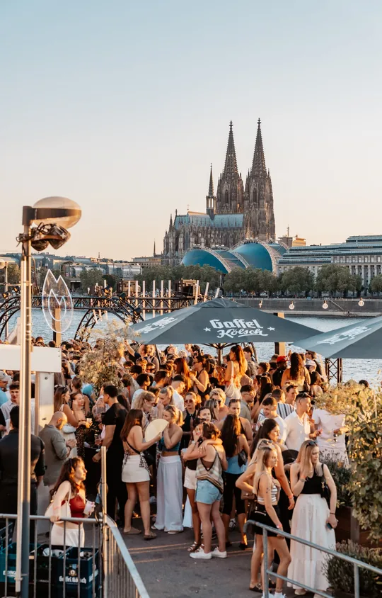 Golden Flame Open Air an den Rheinterrassen Köln: volle Terrasse im Abendlicht, dahinter Rhein, Hohenzollernbrücke und Dom