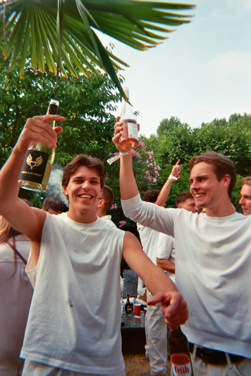 Zwei Freunde in Weiß heben Flasche und Becher in die Sonne