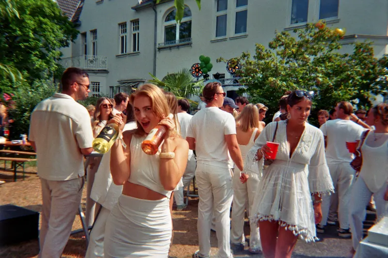 Gäste in weißen Outfits in einem Garten, eine Frau mit Flasche in der Hand lacht in die Kamera