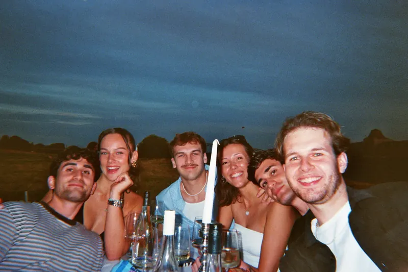 Sechs Freunde sitzen in der Dämmerung an einer langen Tafel mit Kerzen und Weingläsern